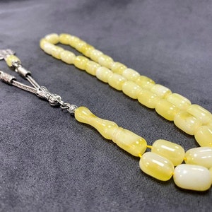 Puede incluir: Un collar de cuentas de oraci&oacute;n de &aacute;mbar amarillo con detalles plateados. Las cuentas son de forma ovalada y tienen un acabado liso y pulido.