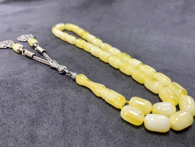 Puede incluir: Un collar de cuentas de oraci&oacute;n de &aacute;mbar amarillo con detalles plateados. Las cuentas son de forma ovalada y tienen un acabado liso y pulido.