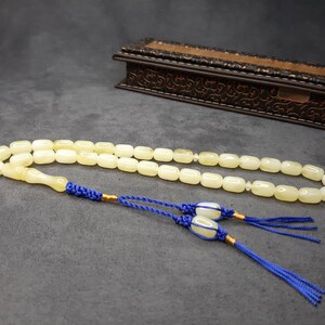 May include: A white prayer beads necklace with a tassel made of blue cord. The beads are oval shaped and the tassel is tied to the end of the necklace.