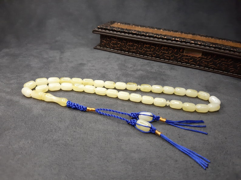 May include: A white prayer beads necklace with a tassel made of blue cord. The beads are oval shaped and the tassel is tied to the end of the necklace.