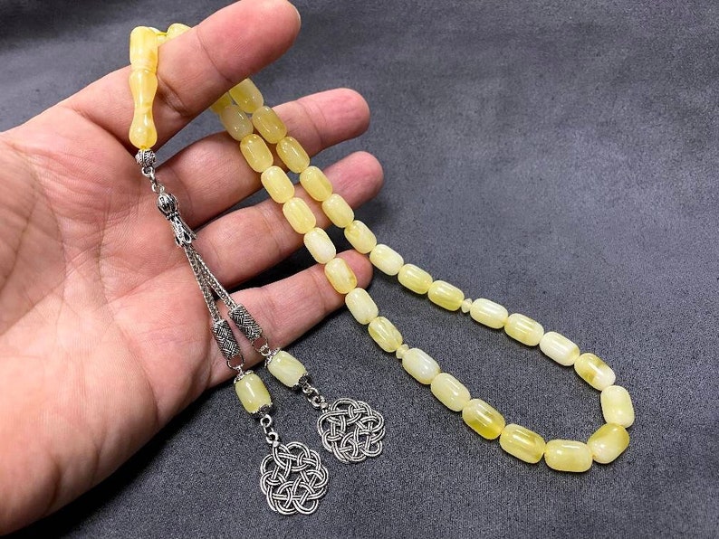 Puede incluir: Un collar de cuentas de oraci&oacute;n de &aacute;mbar amarillo con detalles plateados y un borla. Las cuentas son de forma rectangular y tienen un acabado liso y pulido.