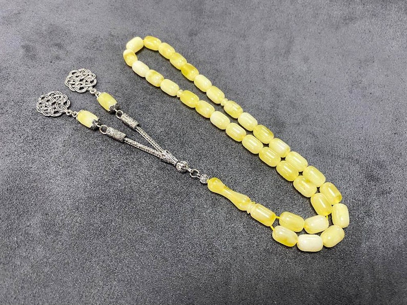 Puede incluir: Un collar de cuentas de oraci&oacute;n de &aacute;mbar amarillo con detalles plateados. Las cuentas son de forma ovalada y el collar tiene un borla.