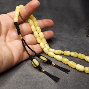 May include: A yellow amber prayer beads necklace with black tassels and gold accents. The beads are oval shaped and strung on a black cord.