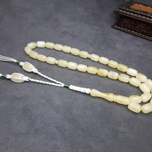 May include: A white prayer beads necklace with a tassel. The beads are oval shaped and have a slight yellow tint. The tassel is made of white cord with green accents.