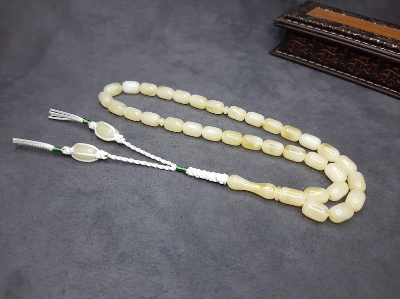 May include: A white prayer beads necklace with a tassel. The beads are oval shaped and have a slight yellow tint. The tassel is made of white cord with green accents.