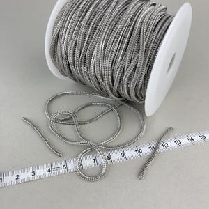 Può includere: Un rotolo di cordoncino metallico argentato con una trama intrecciata. Il cordoncino è avvolto attorno a un rotolo bianco e c'è una lunghezza di cordoncino che giace accanto al rotolo. Un righello è nell'immagine con segni da 1 a 15.