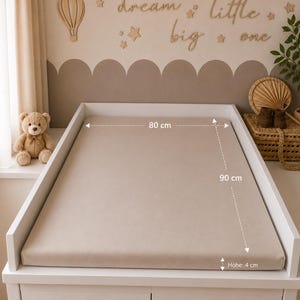 May include: A beige changing pad, 90 cm long, 80 cm wide, and 4 cm high, sits on a white changing table. A teddy bear and decorative wall art with the words "dream little big one" are in the background. The room has a neutral color scheme.