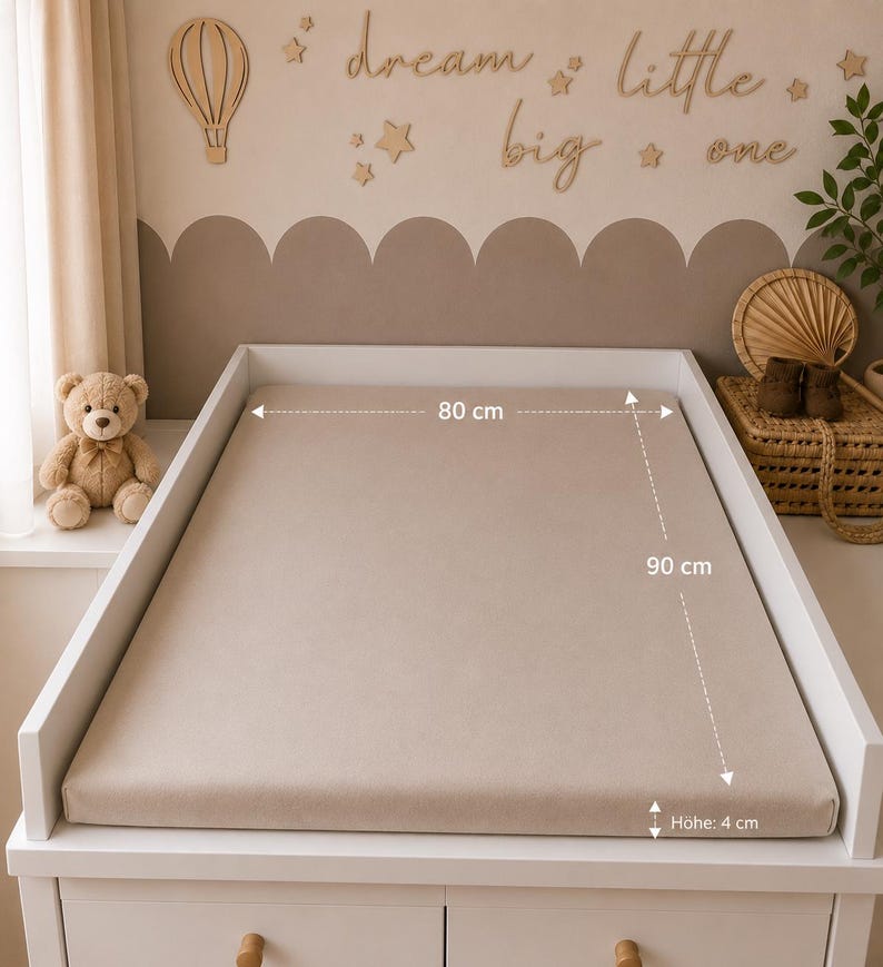 May include: A beige changing pad, 90 cm long, 80 cm wide, and 4 cm high, sits on a white changing table. A teddy bear and decorative wall art with the words "dream little big one" are in the background. The room has a neutral color scheme.