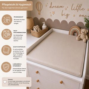 May include: A beige changing pad sits atop a white dresser with wooden knobs. The pad is surrounded by decorative items, including a wooden rainbow, a teddy bear, and a woven basket. The wall features wooden cutouts that spell out "dream little big one."