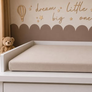 May include: A white changing table with a beige changing pad. The wall behind the table has a brown scalloped design and wooden cutouts of a hot air balloon, stars, and the words "dream little big one."