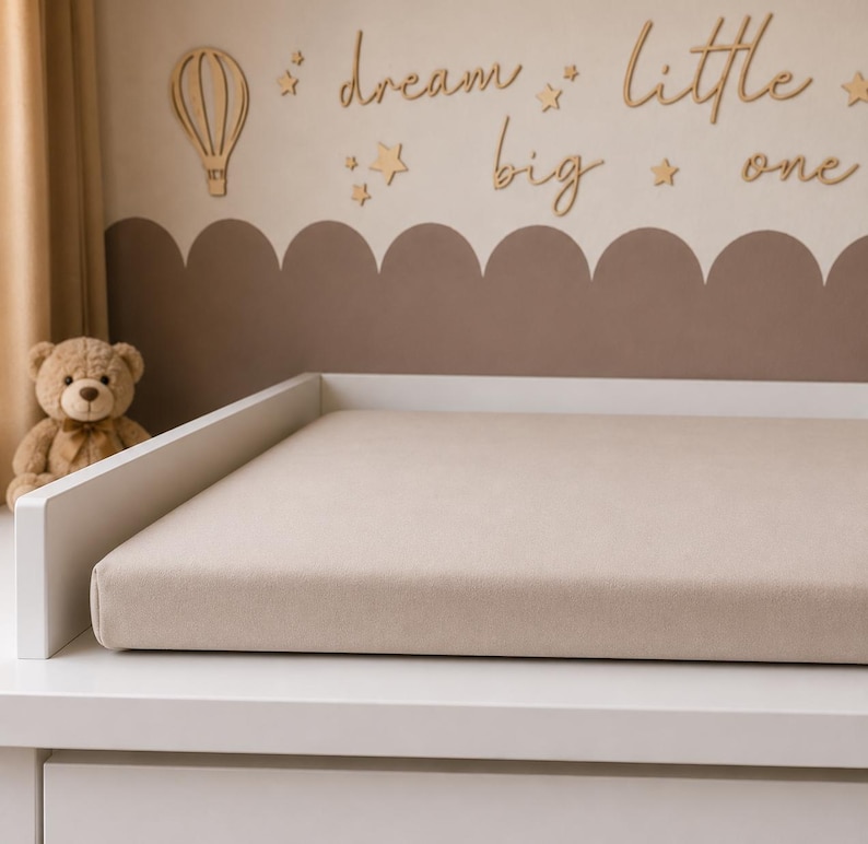 May include: A white changing table with a beige changing pad. The wall behind the table has a brown scalloped design and wooden cutouts of a hot air balloon, stars, and the words "dream little big one."