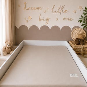 May include: A neutral-toned nursery scene with a crib featuring a beige fitted sheet. The wall is decorated with wooden stars and the phrase "dream little big one." A teddy bear sits on the windowsill, and a woven basket holds baby shoes. The overall aesthetic is soft and calming.