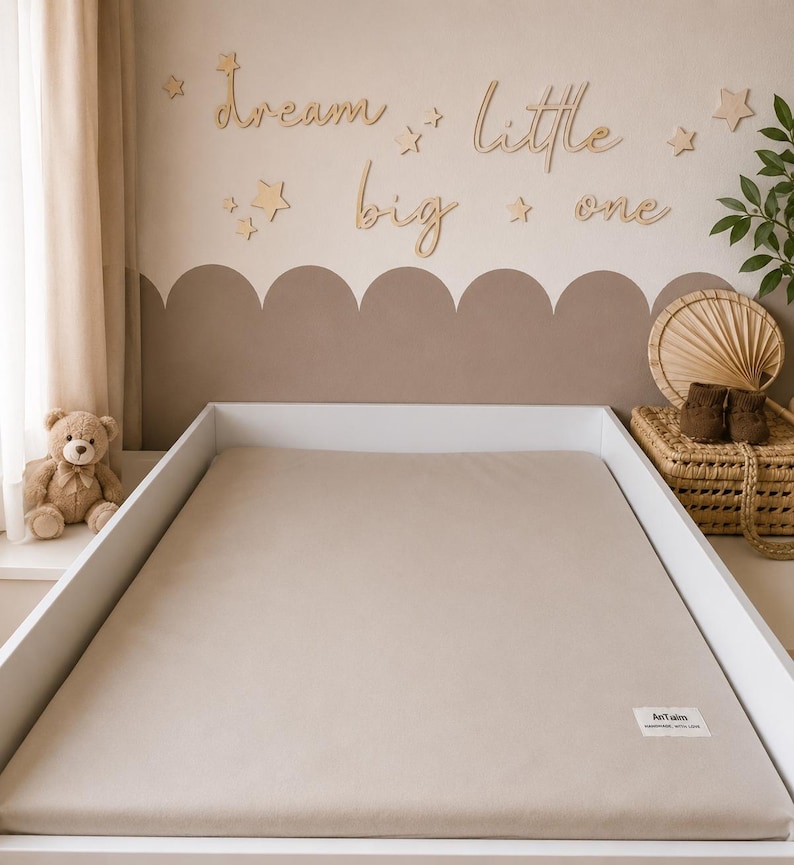 May include: A neutral-toned nursery scene with a crib featuring a beige fitted sheet. The wall is decorated with wooden stars and the phrase "dream little big one." A teddy bear sits on the windowsill, and a woven basket holds baby shoes. The overall aesthetic is soft and calming.