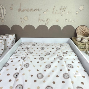 Könnte beinhalten: Weißer Wickelauflagenbezug mit einem braunen und beigefarbenen Löwenmuster. Die Wickelauflage liegt auf einem weißen Wickeltisch. Die Wand hinter dem Wickeltisch ist hellbraun und hat Holzausschnitte mit der Aufschrift "dream big little one".