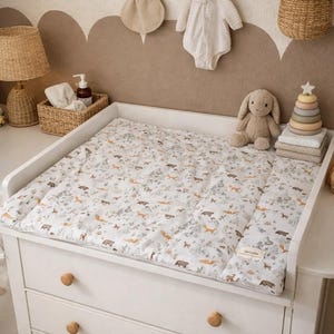 Könnte beinhalten: Weißer Wickeltisch mit einer gemusterten Wickelauflage mit Waldtieren. Ein Plüschhase, ein Holzstapelspielzeug und ein Korb mit Babyausstattung befinden sich auf dem Tisch. Eine Lampe und Wanddekoration vervollständigen die Einrichtung des Kinderzimmers.