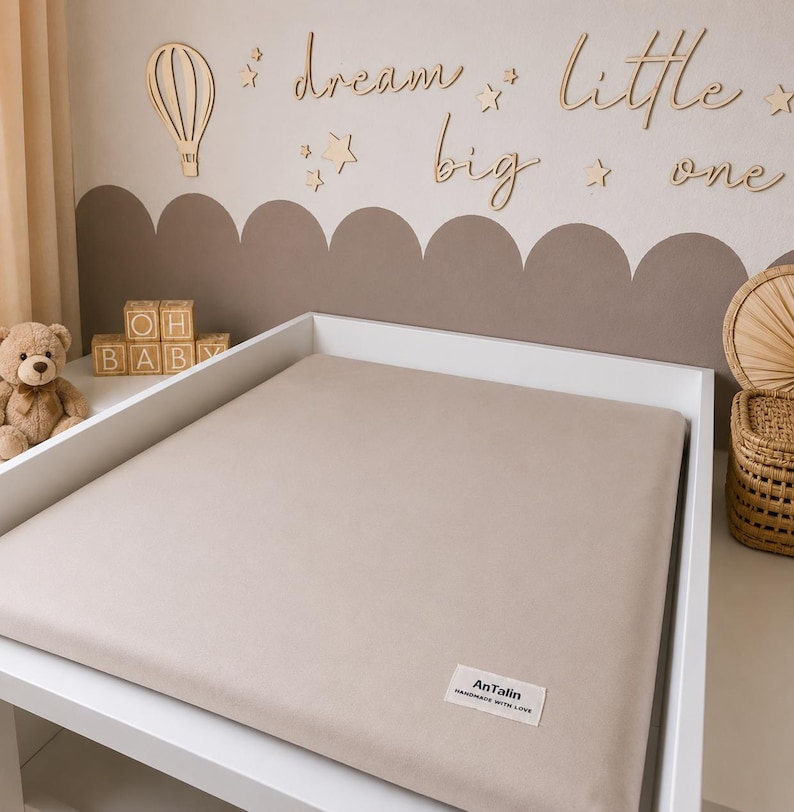 May include: A beige changing pad cover on a white changing table. The cover has a soft, plush appearance. The background features wooden wall decor with the words "dream big little one" and a hot air balloon.