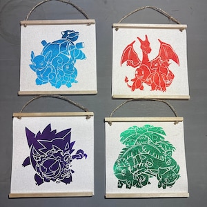 May include: Four framed canvas prints featuring different Pokemon characters. The prints are in a variety of colors, including blue, red, green, and purple. Each print features a different Pokemon character, including a turtle, a dragon, a plant, and a ghost.