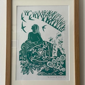 May include: A framed green and white print depicting a woman looking up at birds flying above trees. The print includes rabbits, flowers, and the text "The Seamstress of Spring Tilly". The frame is made of light brown wood.