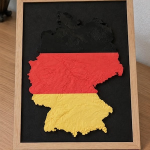 Könnte beinhalten: Ein gerahmtes Wandbild mit einer dreidimensionalen Reliefkarte von Deutschland. Die Karte ist in horizontalen Streifen in Schwarz, Rot und Gold gehalten und befindet sich vor einem schwarzen Hintergrund in einem hellen Holzrahmen.