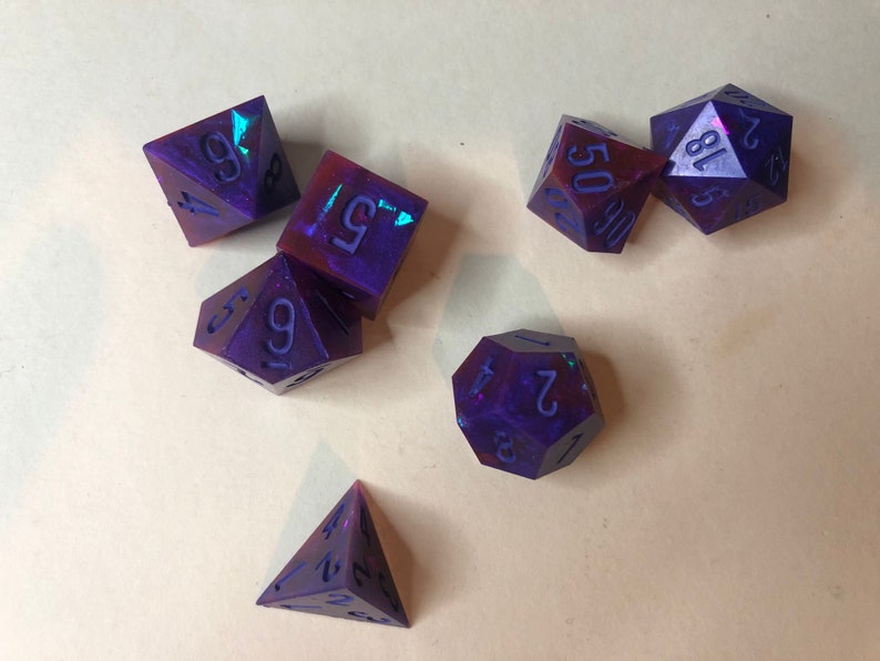 May include: A set of six iridescent purple and red dice with black numbers. The dice are a d4, d6, d8, d10, d12, and d20.