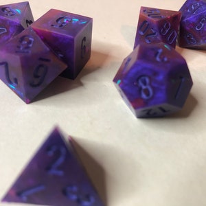 May include: A set of six iridescent purple and pink dice with black numbers. The dice are in various shapes, including a d4, d6, d8, d10, d12, and d20.