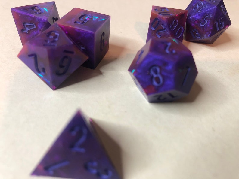May include: A set of six iridescent purple and pink dice with black numbers. The dice are in various shapes, including a d4, d6, d8, d10, d12, and d20.