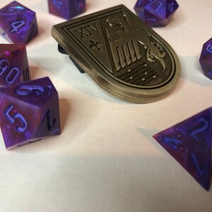 May include: A set of purple and blue iridescent dice with a gold metal emblem featuring a stylized bird and the Roman numeral XV. The dice are a variety of shapes and sizes, including a d4, d6, d8, d10, d12, and a d20.