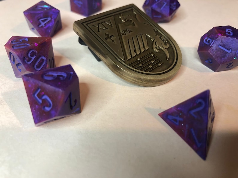 May include: A set of purple and blue iridescent dice with a gold metal emblem featuring a stylized bird and the Roman numeral XV. The dice are a variety of shapes and sizes, including a d4, d6, d8, d10, d12, and a d20.