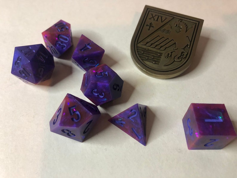 May include: A set of seven iridescent dice with purple and pink swirls. The dice are numbered 1 through 6 and are made of a translucent material. The dice are scattered on a white surface.