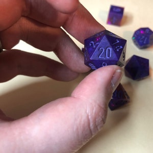 May include: A set of five purple iridescent dice with black numbers. The dice are a D20, D12, D10, D8, and D6.