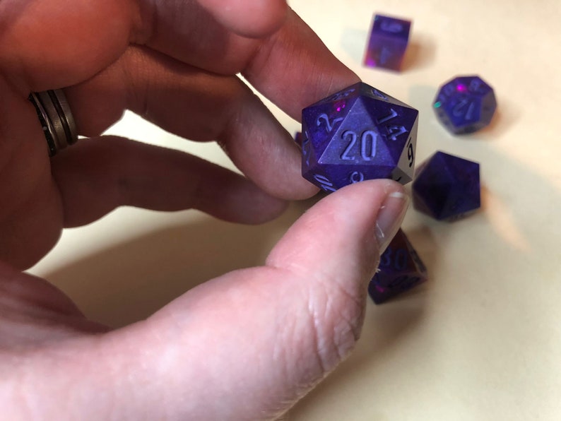 May include: A set of five purple iridescent dice with black numbers. The dice are a D20, D12, D10, D8, and D6.