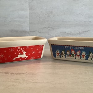 May include: Two ceramic loaf pans with festive holiday designs. One pan features a red background with white snowflakes and a white reindeer. The other pan has a blue background with white snowmen and the text "LET IT SNOW".