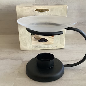 May include: A black metal oil warmer with a frosted glass dish. The warmer has a curved handle and a base for a tea light. The box behind it reads "Florasense" and mentions a simmer vase.