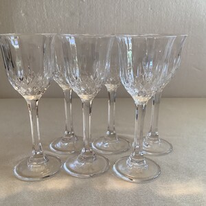 May include: Set of six clear glass wine glasses with a cut crystal design. The glasses are arranged in a semi-circle on a white surface.