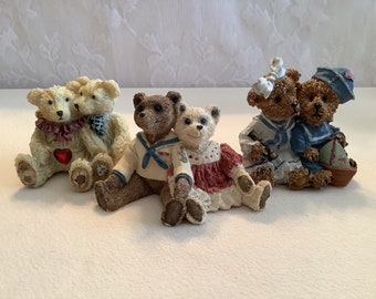 May include: Four pairs of decorative teddy bear figurines. The bears are in various outfits, including sailor suits and dresses. Some bears are hugging, and one pair has a heart on its chest. The figurines are in shades of brown, cream, red, and blue.