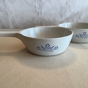 May include: Two white ceramic bowls with blue floral designs. The bowls have a handle on one side and are perfect for serving soup or other dishes.