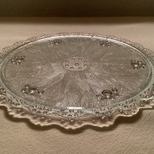 May include: A clear glass cake plate with a scalloped edge and a raised, textured floral design in the center. The plate has a beaded edge.