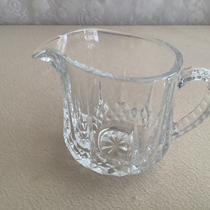 May include: A clear glass creamer with a diamond pattern and a handle. The creamer is empty and sitting on a light brown surface.
