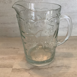 May include: A clear glass pitcher with a floral design. The pitcher has a handle and a wide mouth.