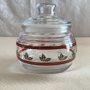 May include: A clear glass jar with a lid, decorated with a red and green holly pattern. The jar has a textured surface and is perfect for storing holiday treats.