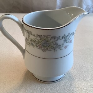 May include: White porcelain creamer with a floral design in shades of pink, blue, and green. The creamer has a gold trim around the top and bottom.