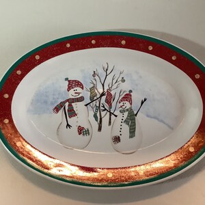 May include: Oval white ceramic serving platter with a green and red border. The platter features a winter scene with two snowmen and a tree. The border has gold glitter accents.