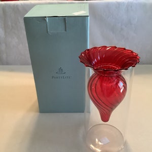 May include: A red glass votive holder with a swirl design, in a clear glass cylinder, with a PartyLite box in the background.