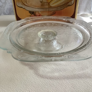May include: A clear glass cake stand with a decorative etched design. The stand has a square base and a round top with a center pedestal.