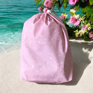 Puede incluir: Una bolsa de cordón rosa con un patrón de estrellas blancas. La bolsa está sentada en una playa de arena blanca.