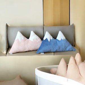 以下が含まれることがあります： 山型の装飾用クッション2つ。1つは白い雪のピンク色、もう1つは白い雪の青色です。灰色のベンチに置かれています。白い容器には、追加の山型クッションが見えます。