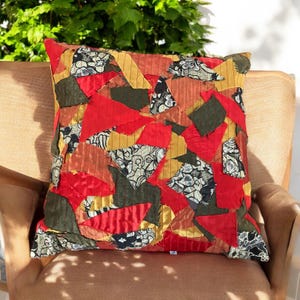 May include: A square throw pillow with a patchwork design. The pillow is made of various fabrics in shades of red, gold, and green. The fabrics are cut into different shapes and sewn together to create a unique and abstract pattern.