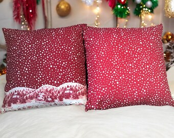 Op de afbeelding: Twee vierkante decoratieve kussens. De kussens zijn dieprood met een wit stippenpatroon. Eén kussen heeft een winterlandschap aan de onderkant. De kussens liggen op een witte ondergrond.