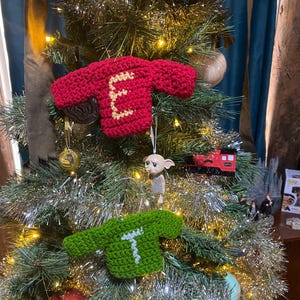 Peut inclure: Un sapin de Noël décoré avec des guirlandes et des ornements, dont deux décorations de pull en crochet faites à la main. L'une est rouge avec un "E" crème, l'autre est verte avec un "T" blanc. Autres décorations.