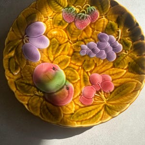 Può includere: Un piatto in ceramica giallo con un design a rilievo di frutta, tra cui uva, fragole e ciliegie. Il piatto ha un aspetto rustico e vintage.